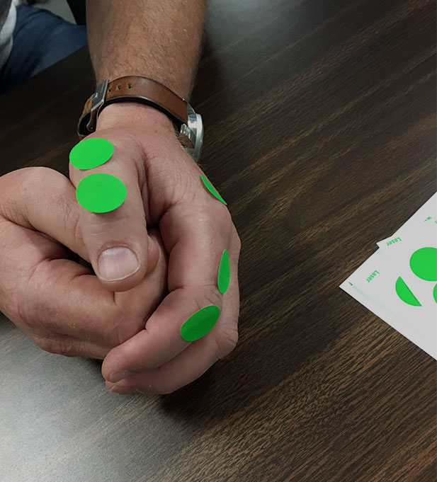 Two hands clasped together with green dot stickers on the fingers after completing a Make-a-card Lean simulation.