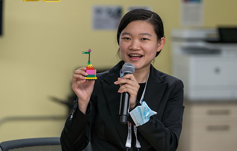 A woman holds a microphone and a small tower built out of LEGOs, which was created during a Lean simulation.