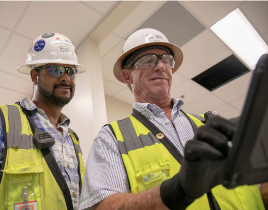 Two men in yellow vests, white hard hats, and safety glasses smile while looking at an iPad stating their Lean mission.