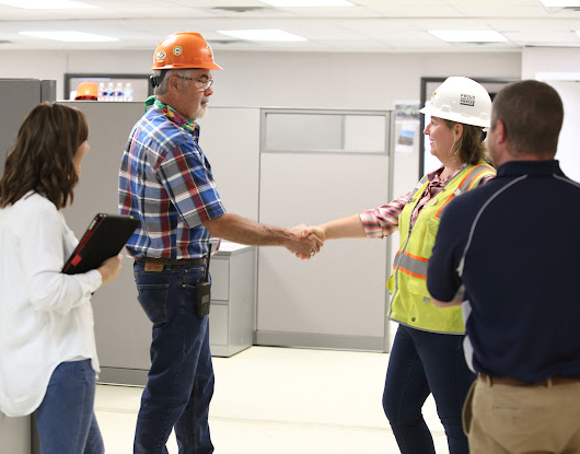 A man in an orange hard hat and glasses shakes hands with a woman in a yellow vest and white hard hat inside an office.